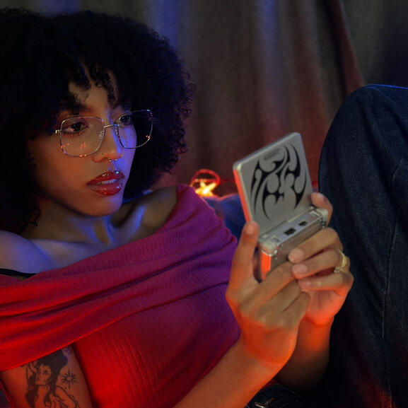 Mixed Black girl with curly afro, gold glasses, and a tattoo on her arm of a mermaid surrounded by stars (representing the zodiac sign Aquarius) is wearing a red off-the-shoulder top, blue baggy jeans and holding a retro Gameboy. There are red star-shaped