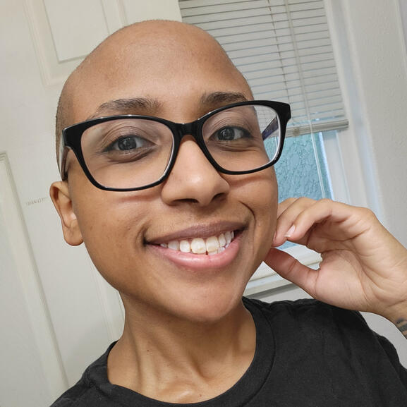 AmethystMoon smiling at the camera. She has a bald head and is wearing glasses.