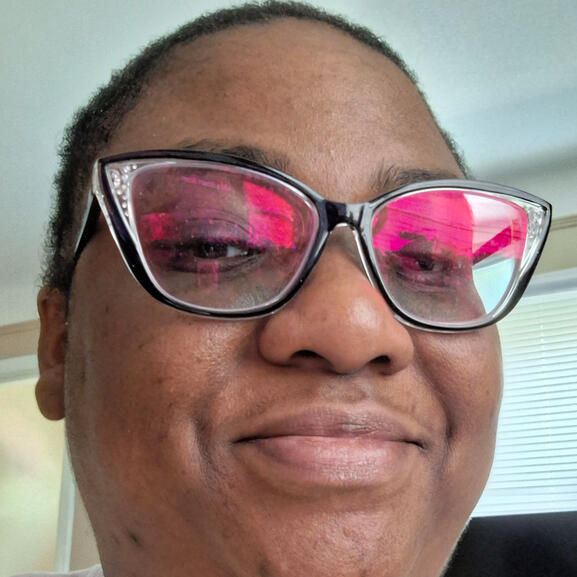 Darunia, a Black non-binary person with black glasses and short hair, is smiling at the camera.