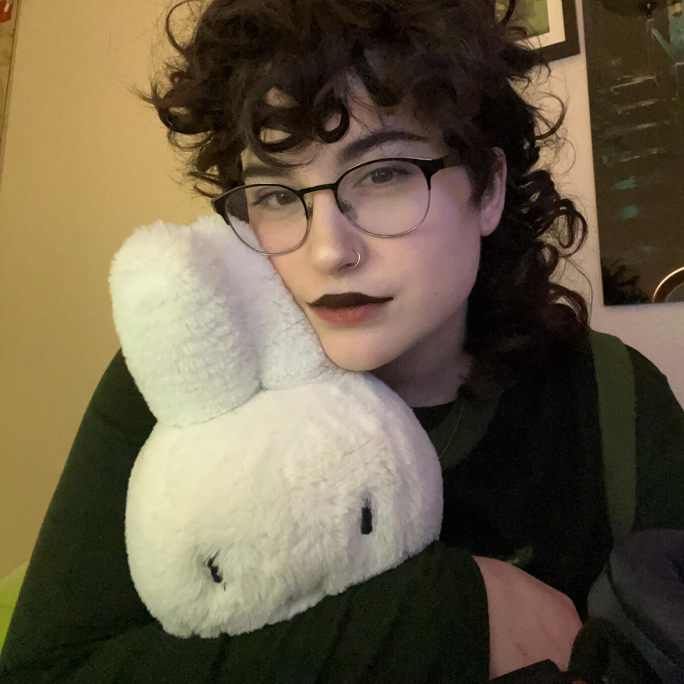 Jade, a white person with brown curly hair is holding a stuffed Miffy rabbit close to their face. They are wearing large glasses and have dark lipstick on just their upper lip. They are wearing a dark sweater vest with green trim, and are in front of an of