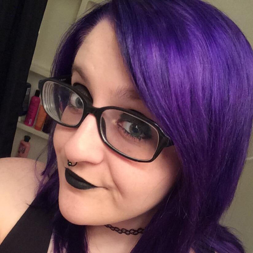 A closeup/selfie of a white person with purple hair. They are wearing black glasses, a black septum ring, a black tattoo-style choker, black lipstick and black eye shadow. They are wearing a black tank top and on their chest there is a tattoo of a heart wi