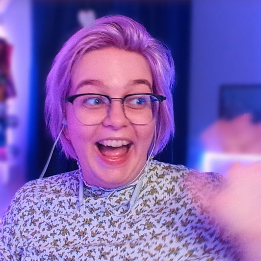 A woman with short pink hair smiling widely, while waving excitedly at the camera. She is wearing glasses with a black top rim, and a floral top with a high neck. The background is softly blurred and in purple and pink colors, creating a joyful and vibrant