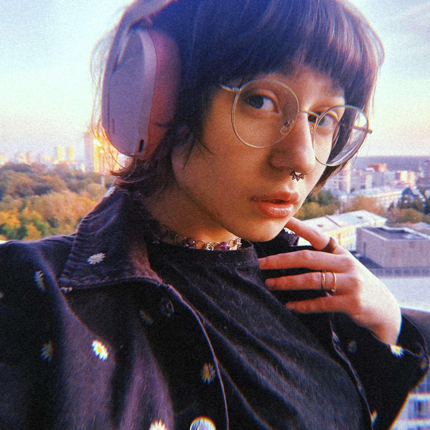 a white femme-presenting person is standing on a balcony with a city landscape in the background. they have a brown bob with bangs, hazel eyes, a big nose and a septum piercing. they're wearing glasses, headphones, rings, a necklace, a plain black t-shirt