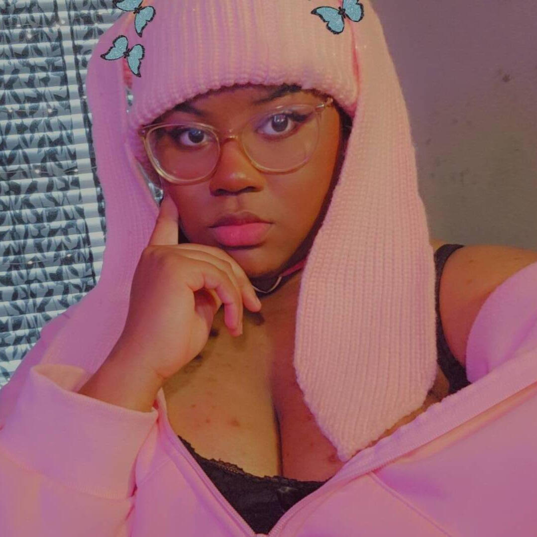 Black girl with clear square glasses and a light pink beanie with long rabbit ears and a pink half zipped hoodie with a black tank top underneath, and her hand on the side of her face.
