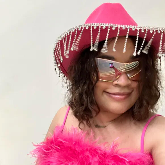 A selfie of Bri (she/they), wearing a pink cowboy hat with rhinestones and a fuzzy pink top.