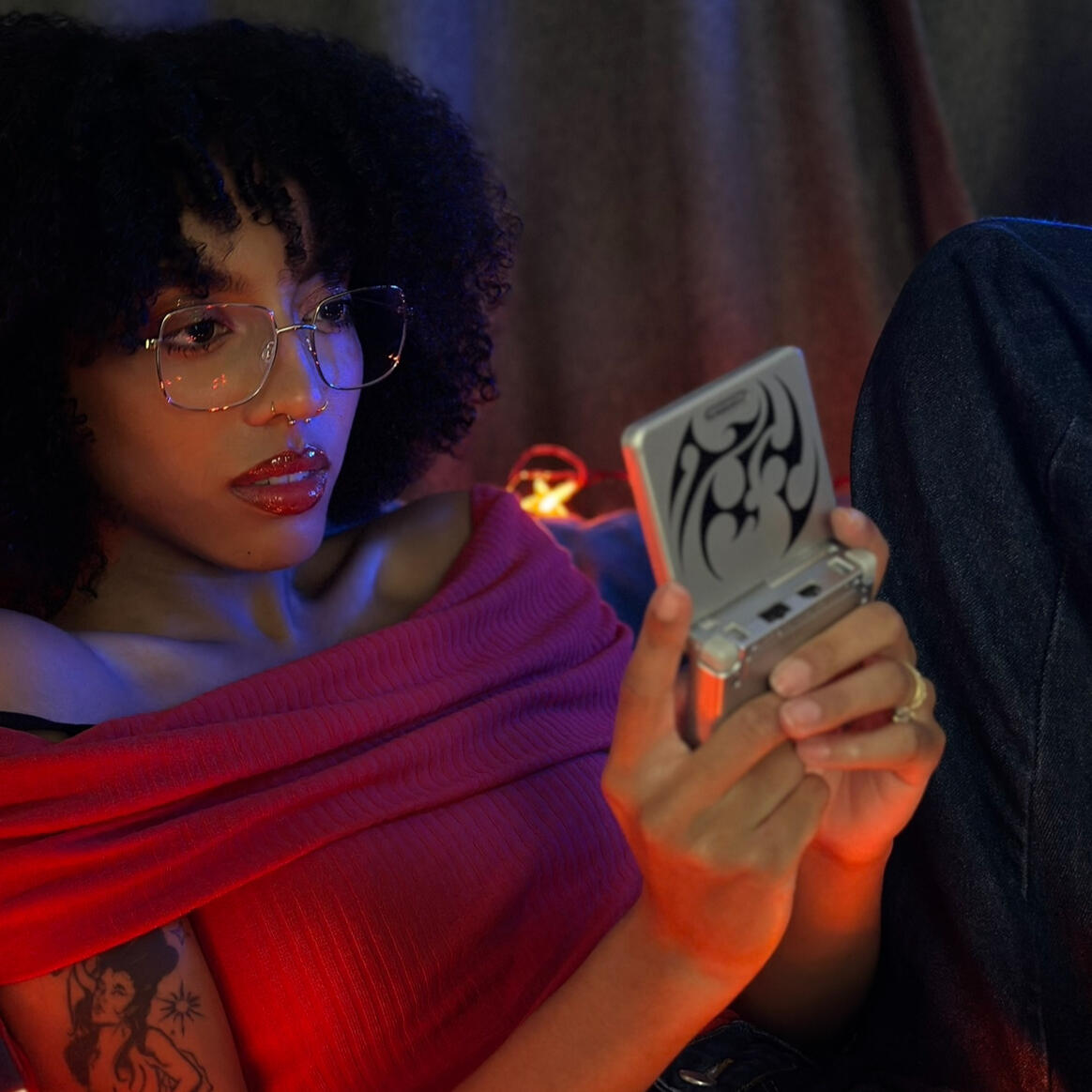 Mixed Black girl with curly afro, gold glasses, and a tattoo on her arm of a mermaid surrounded by stars (representing the zodiac sign Aquarius) is wearing a red off-the-shoulder top, blue baggy jeans and holding a retro Gameboy. There are red star-shaped