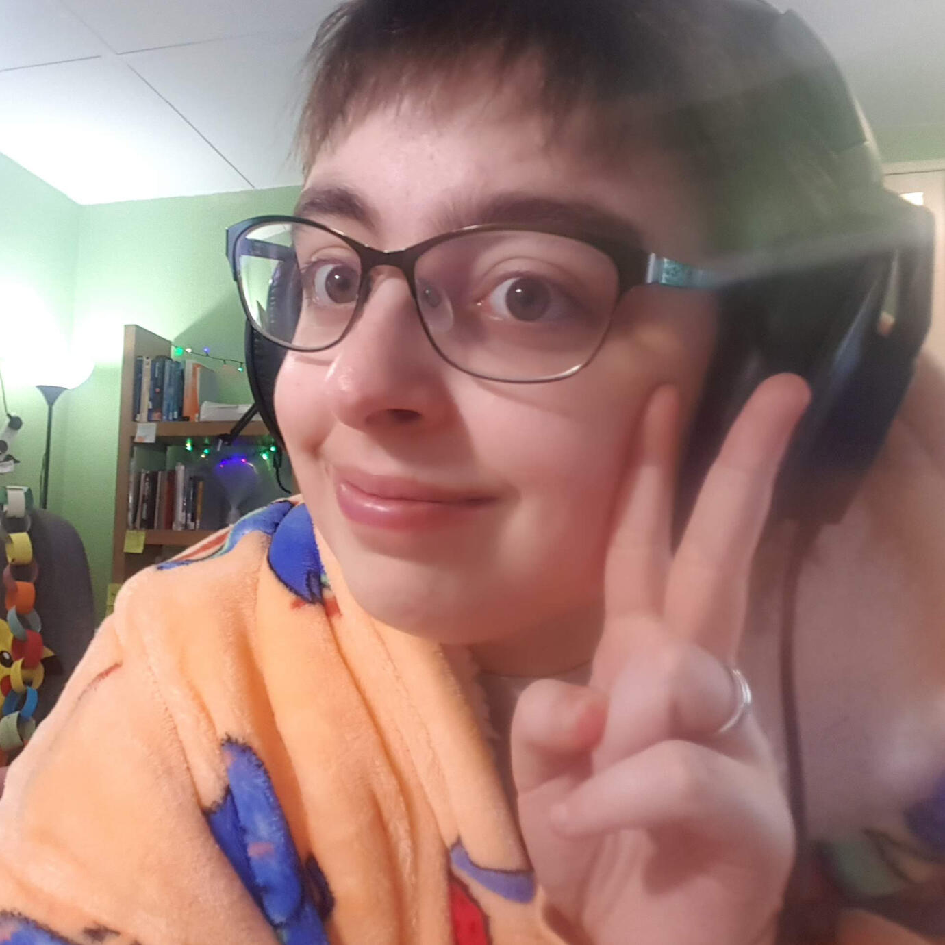A photo of Artie, a white person with short brown hair and glasses, smiling at the camera doing a peace sign with their fingers.