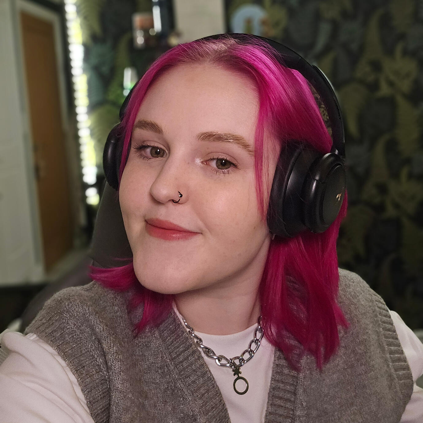 Lozza, a white person with pink hair wearing black headphones smiling at the camera. They're wearing a chain necklace with the non-binary gender symbol on it, and there are lights and leafy wallpaper in the background which is slightly blurred