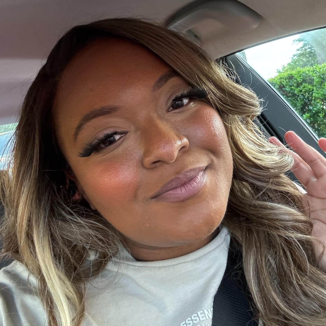 Kayluhh, a woman with long wavy blonde-highlighted hair, smiling in a car while wearing a beige shirt. The photo shows her warm, confident personality