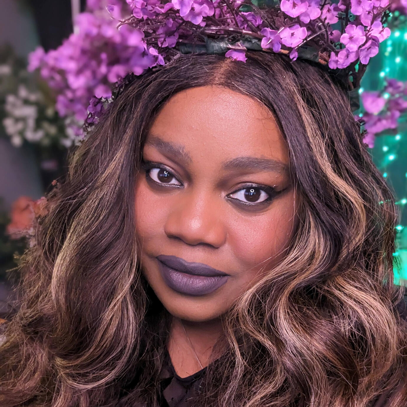 Picture of Marcy with long, wavy brown and blonde-highlighted hair poses in front of a background filled with purple flowers and soft green lights. They wear a crown made of small purple blossoms and have a slight smiling expression with bold dark lipstick