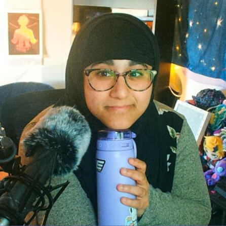 a photo of omni, accompanied by their microphone and giving a small smile. they are wearing a black hijab, and a beige sweater whose bodice is black with floral designs. they are holding a light purple water bottle and resting their chin on it.