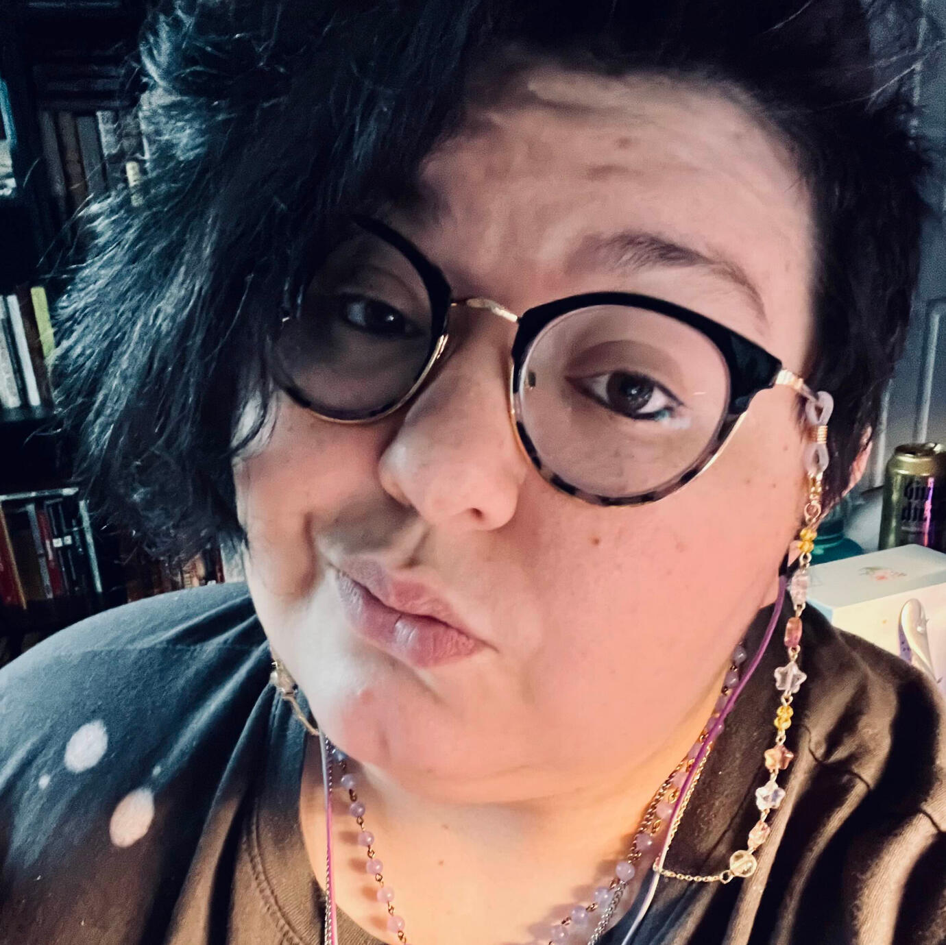 A selfie of quinn. They have short brown hair and wear cat eye black and gold glasses with an attached glasses chain of stars, a gray shirt, and a purple beaded necklace. They smirk at the camera.