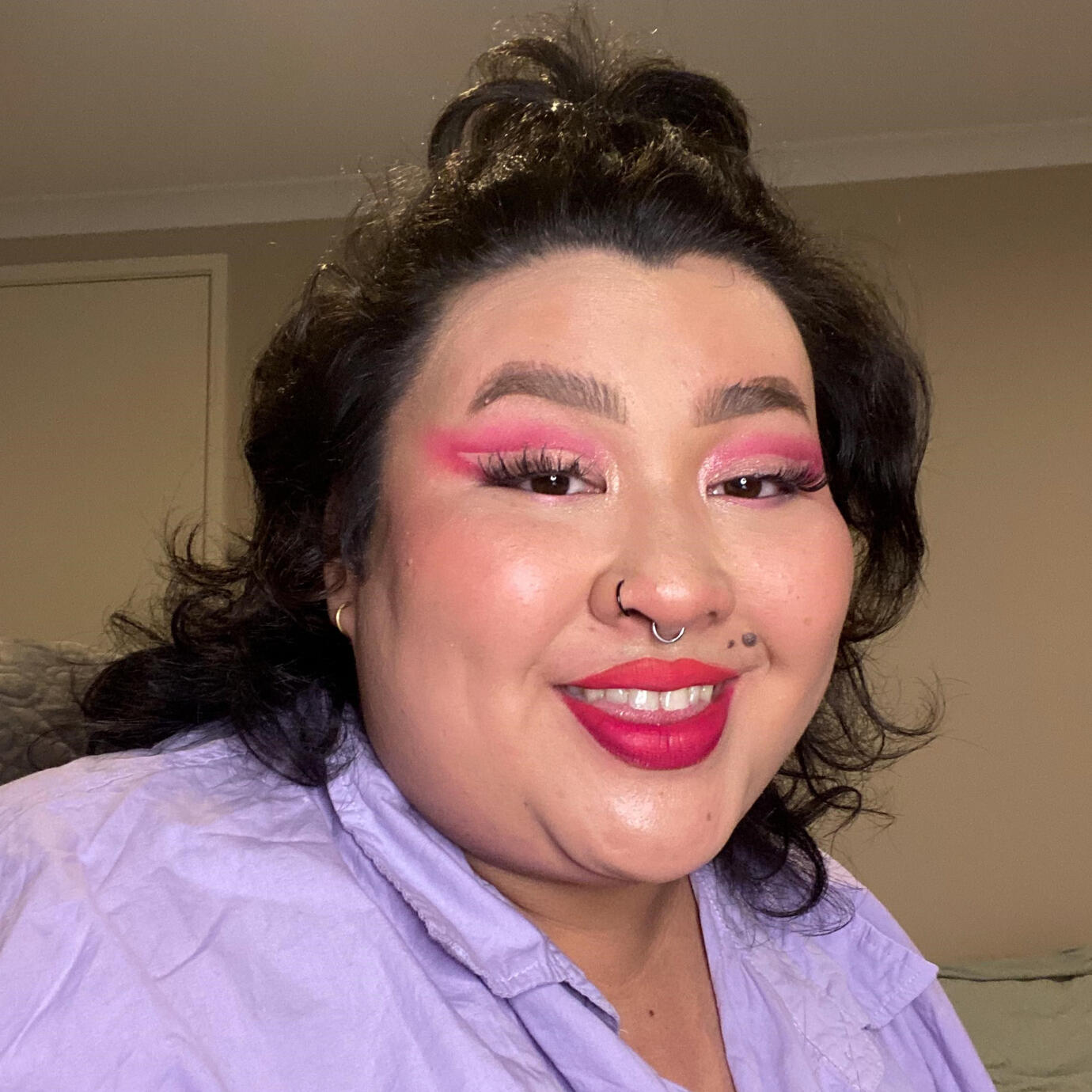 A selfie of Clore, a fat, fair-skinned Asian person with dark brown hair and eyes, with a slight smile. They are wearing a lavender button down shirt. They have a pink cut-crease eyeshadow look with wispy false lashes. Their hair is in a half-up and hal