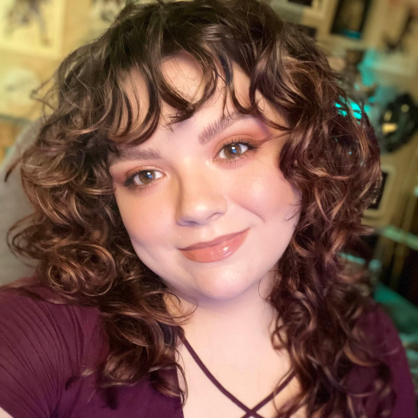 Zen (she/her) is indoors and wearing a maroon top with chest detail and has red curly hair with dimensional highlights. She is wearing red-brown toned makeup that compliments her very pale skin and large brown eyes. She has a closed-lip smile and looks hap