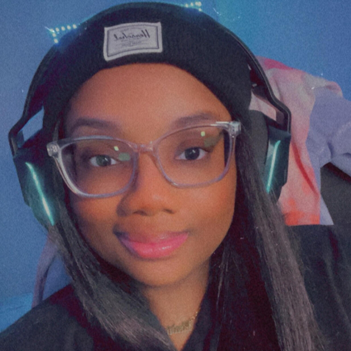 A selfie of Mira (they/them), a Black, nonbinary femme person. Mira has brown skin and medium length dark brown straight hair. They are wearing a black t-shirt, a black knit beanie, and clear framed glasses and a gold script necklace that reads "radical".