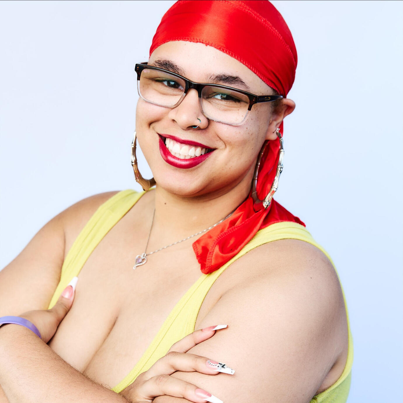 Autumn is smiling wearing glasses with a red durag. She is wearing a yellow tank top and is crossing her arms looking at the camera.