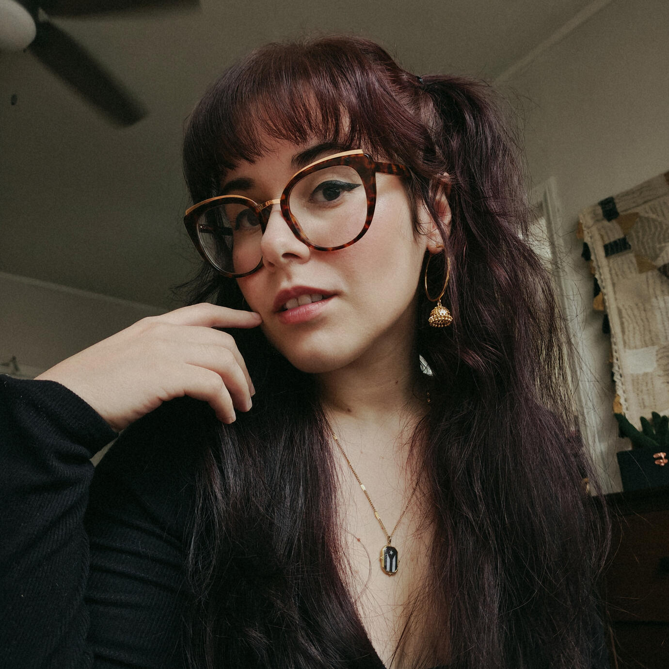 selfie of Andrea wearing a black long sleeved top. her hand is resting by her face. her hair is half up and long. she has gold hoops on, makeup on and glasses on and is looking at the camera slightly smirking with her head tilted.