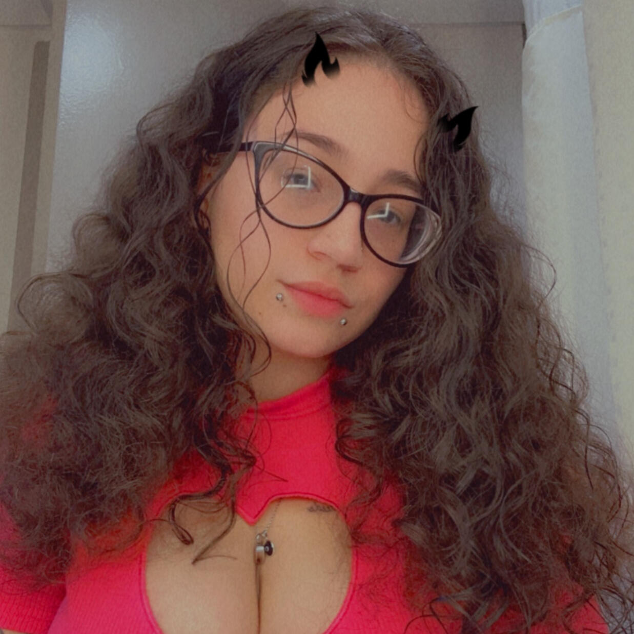 Image is a selfie of Beersnack_. They have long, curly brown hair. They are wearing black glasses, silver snake bite piercings, a silver necklace, and a pink shirt with a heart shaped cut-out on the chest. They have one arm extended to hold the camera and
